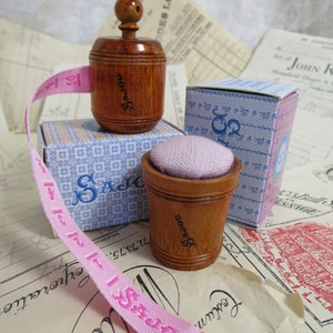 May include: A pink fabric measuring tape with a wooden spool and a small wooden thimble with a pink cushion. The spool and thimble are labeled "SAJOU".