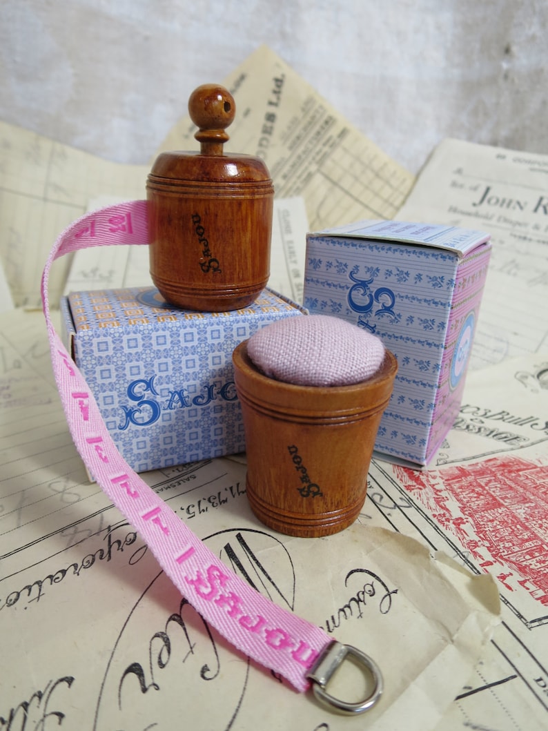 May include: A pink fabric measuring tape with a wooden spool and a small wooden thimble with a pink cushion. The spool and thimble are labeled "SAJOU".