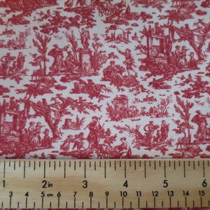 Op de afbeelding: Rode toile stof met een herhalend patroon van gestileerde bomen, huizen en figuren. De stof wordt gemeten met een liniaal die 5 cm aangeeft.