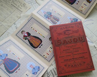 Peut inclure: Un ensemble de patrons de point de croix ou sont représentées des figures en costumes traditionnels, avec un livre rouge intitulé "Album de Point de Croix" par Maison Sajou. Les motifs sont sur fond beige, avec diverses couleurs.