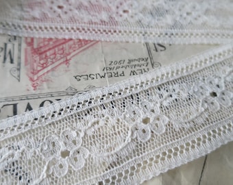 May include: White lace trim with a delicate floral pattern. The lace is folded over itself and is lying on a piece of paper with text that reads "New Premises Established 1951 Redhill 1902".