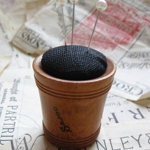 May include: A wooden pincushion with a black fabric top and four white pearl-headed pins. The pincushion is sitting on a piece of paper with text.