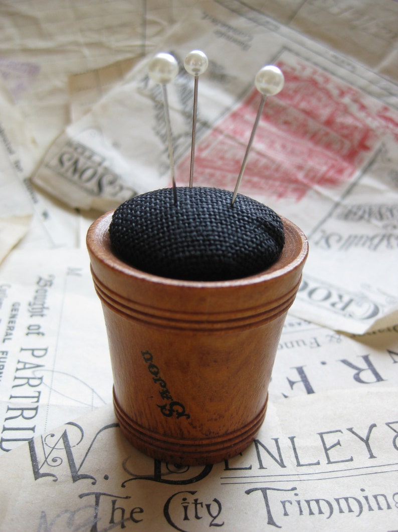 May include: A wooden pincushion with a black fabric top and four white pearl-headed pins. The pincushion is sitting on a piece of paper with text.