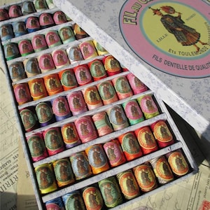 May include: A vintage box of Fil au Chinois embroidery thread spools. The box is pale blue with a floral pattern and a pink circle with the text "Fil au Chinois" in black. The spools are arranged in rows and are a variety of colours, including red, orange, yellow, green, blue, and purple.