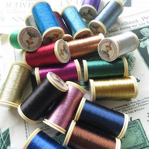 May include: A collection of spools of metallic thread in various colors, including gold, silver, red, blue, green, and purple. The spools are labeled with the text "FILS A COUDRE CLASSIQUES ET FANTAISIE AU CHINOIS".