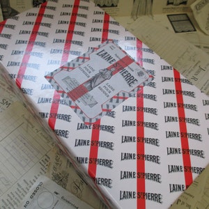 May include: A white gift box with red stripes and the text "LAINE ST PIERRE" repeated across the surface. The box has a smaller label with the text "LAINE ST PIERRE" and the words "LAINE POUR REPRISER" and "LAINE POUR BRODER" in French.