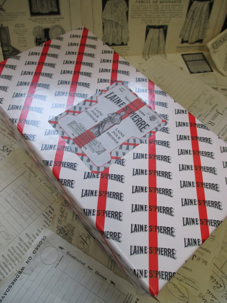 May include: A white gift box with red stripes and the text "LAINE ST PIERRE" repeated across the surface. The box has a smaller label with the text "LAINE ST PIERRE" and the words "LAINE POUR REPRISER" and "LAINE POUR BRODER" in French.