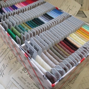 May include: A white box filled with spools of embroidery floss in various colors. The box is labeled "J. & J.  Lud.  Ltd."