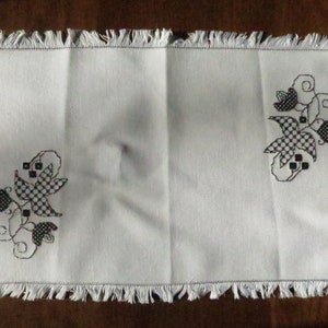 Puede incluir: Un camino de mesa blanco con un diseño floral bordado en blanco y negro. El camino de mesa tiene un borde con flecos.