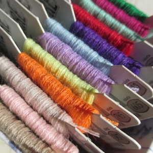 May include: A close-up of a variety of embroidery floss spools in a white plastic organizer. The spools are arranged in rows and are labeled with numbers. The floss colors include pink, orange, yellow, green, blue, purple, and red.