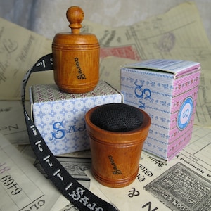 May include: A set of three wooden sewing tools with a black and white patterned box. The tools include a wooden thimble, a wooden pincushion, and a wooden tape measure with a black ribbon. The tape measure has the word "SAJOU" printed on it.