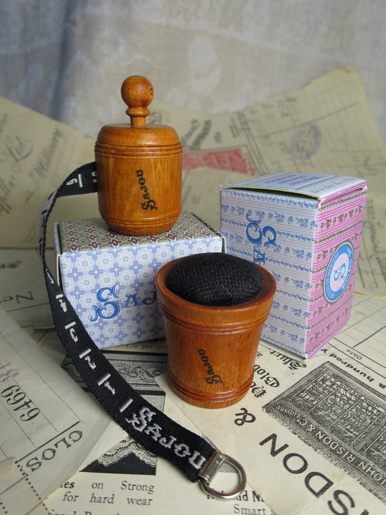 May include: A set of three wooden sewing tools with a black and white patterned box. The tools include a wooden thimble, a wooden pincushion, and a wooden tape measure with a black ribbon. The tape measure has the word "SAJOU" printed on it.