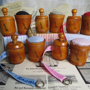 May include: A set of eight wooden pincushions with a vintage look. Each pincushion has a small knob on top and is labeled "SaJou". The pincushions are displayed on a table with a variety of fabrics and ribbons.