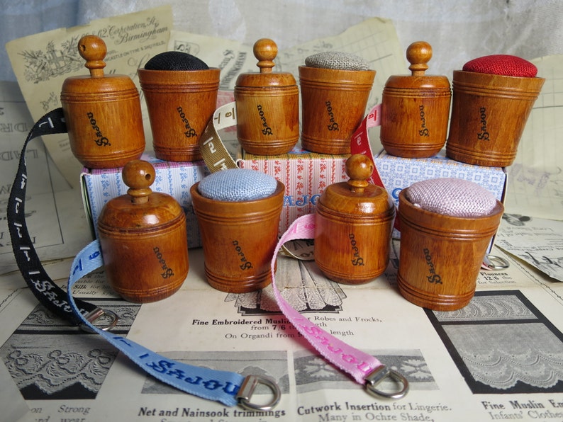 May include: A set of eight wooden pincushions with a vintage look. Each pincushion has a small knob on top and is labeled "SaJou". The pincushions are displayed on a table with a variety of fabrics and ribbons.