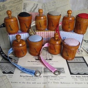 May include: Nine wooden pincushions with different colored fabric tops. The pincushions are arranged in a circle on a table with a vintage fabric pattern. The pincushions are labeled with the word "SAJOU".