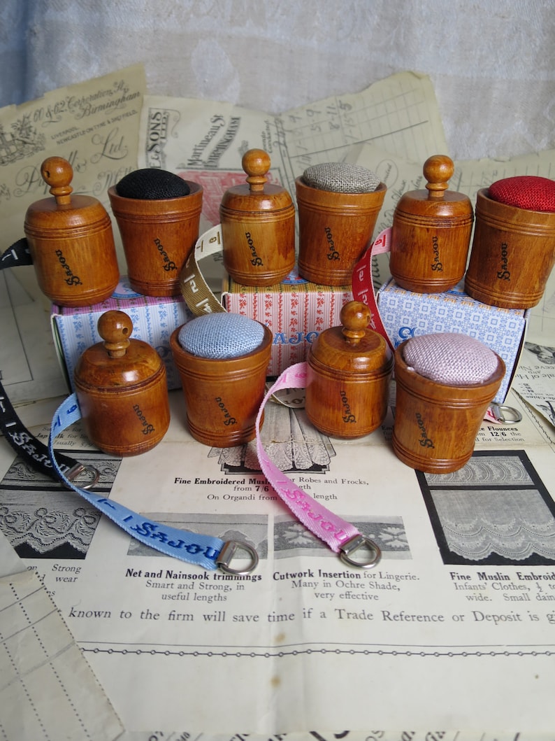 May include: Nine wooden pincushions with different colored fabric tops. The pincushions are arranged in a circle on a table with a vintage fabric pattern. The pincushions are labeled with the word "SAJOU".