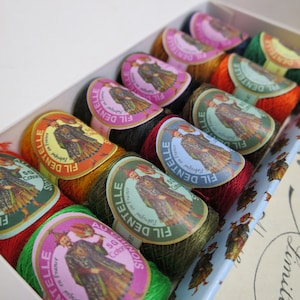 Può includere: Un set di 10 bobine di filo per ricamo in una scatola bianca. Le bobine sono etichettate "Fil Dentelle" e presentano un'illustrazione colorata di una donna in abito tradizionale. Le bobine sono disposte in fila, con colori che vanno dal rosso al verde.
