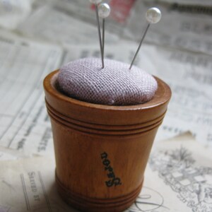 May include: A wooden pincushion with a light purple fabric top and three pearl-headed pins. The pincushion has the word "Dorn" printed on the side.
