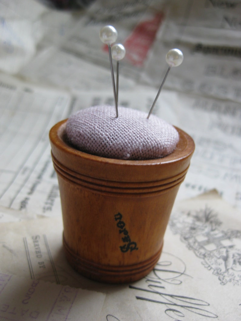 May include: A wooden pincushion with a light purple fabric top and three pearl-headed pins. The pincushion has the word "Dorn" printed on the side.