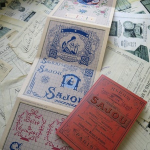 May include: A red book titled "Album de Point de Croix Maison Sajou" with a gold border and decorative flourishes. The book features a variety of embroidery patterns and designs.