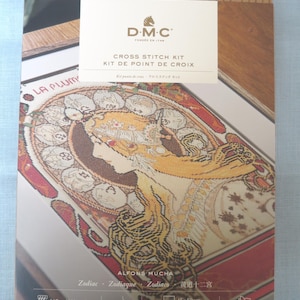 May include: A D.M.C. cross stitch kit featuring an Alfons Mucha Zodiac design. The packaging displays a profile illustration of a person with flowing hair surrounded by zodiac symbols. It includes 165 meters of thread and a 45 by 50 cm fabric piece.