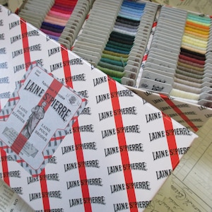 May include: A white box with red stripes and the text "LAINE ST PIERRE" repeated many times. The box is filled with small spools of colorful yarn.