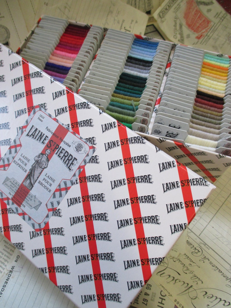May include: A white box with red stripes and the text "LAINE ST PIERRE" repeated many times. The box is filled with small spools of colorful yarn.