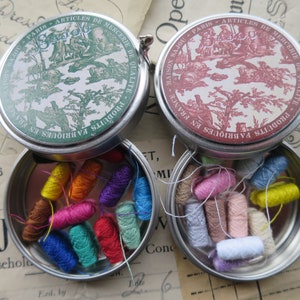 Puede incluir: Dos latas de metal vintage con tapas, cada una contiene carretes de hilo de colores. Las tapas presentan un diseño floral con el texto "Maison Sajou Paris Articles de Mercerie de Qualite Produits Fabriques en France".