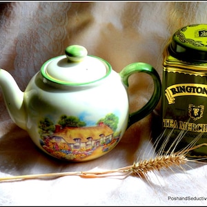 Puede incluir: Una tetera blanca vintage con asa y ribetes verdes, con una escena campestre. Junto a ella, una lata de té Ringtons Tea Merchants verde y dorada. Una espiga de trigo se encuentra frente a la tetera y la lata, sobre una tela color crema.
