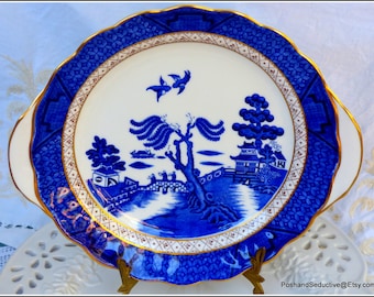 May include: A blue and white porcelain serving plate with a scalloped edge and gold trim. The plate features a traditional willow pattern with a tree, a bridge, and a pagoda.