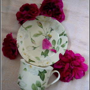 Può includere: Una tazza da tè e un piattino bianchi con un motivo floreale con rose bianche e foglie verdi. La tazza da tè e il piattino sono circondati da rose rosa.