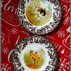 May include: Two decorative plates with dog illustrations. The plates have a white center with a dog scene and a brown floral border. One plate features a beagle, the other two golden retrievers. The plates are on a red background.