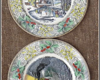 May include: Two vintage ceramic plates with a white background and a green, yellow, and red floral border. The top plate features a winter scene with people ice skating on a frozen lake. The bottom plate features a winter scene with a train traveling through a snowy forest.