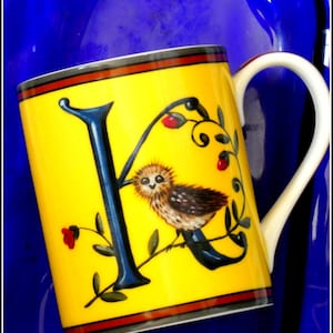 Può includere: Una tazza in ceramica gialla con una grande lettera K blu scuro, un'illustrazione di un piccolo gufo marrone e accenti floreali rossi. La tazza ha un manico bianco e strisce orizzontali rosse e nere sui bordi superiori e inferiori su sfondo blu.