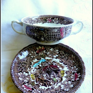 May include: A brown and white ceramic soup bowl with a floral pattern and a scene of people in a town. The bowl is sitting on a matching saucer with a similar floral pattern.