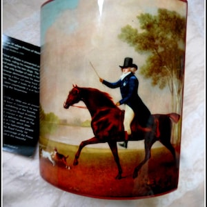 Puede incluir: Un jarrón de cerámica con forma curva y una escena pintada de un hombre a caballo cabalgando por un paisaje. El hombre lleva un sombrero negro y una chaqueta azul. El caballo es marrón y tiene una raya blanca en la cara. El fondo es un cielo azul con nubes blancas y árboles verdes.