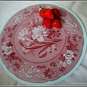 May include: A vintage, pink and white ceramic plate with a floral design. The plate has a raised rim and a scalloped edge. There are five red strawberries on top of the plate.