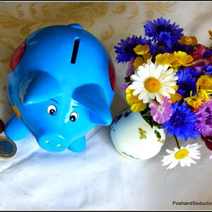 Könnte beinhalten: Ein blaues Sparschwein mit roten und gelben Akzenten steht auf einer weißen Tischdecke mit einem Strauß bunter Blumen in einer weißen Vase.