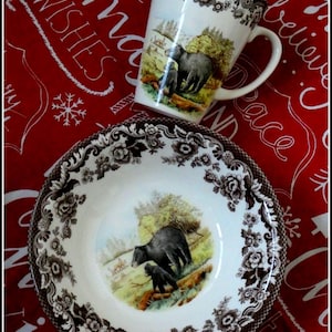 May include: A white ceramic mug and bowl set with a black and brown floral border design. The mug and bowl feature a central image of a bear and cub in a forest scene. The background is a red fabric with white text.