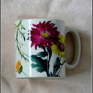 May include: A white ceramic mug featuring a floral design with a large, vibrant magenta flower and a yellow flower. Green leaves and stems accent the flowers. The mug has a curved handle and a glossy finish, perfect for enjoying your favorite beverage.