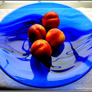 May include: Four ripe peaches on a blue glass serving dish with a wavy design. The peaches are arranged in a cluster in the center of the dish.