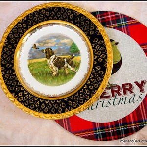 May include: A black and gold decorative plate with a white center featuring a hunting scene with a brown and white dog and a bird. The plate is on top of a red and green plaid placemat with the text "Merry Christmas".