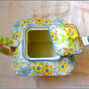 May include: A vintage ceramic teapot with a square shape and a floral pattern in yellow, blue, and orange. The lid is open, revealing the interior. The handle and spout are decorated with flowers.