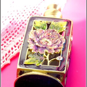 May include: A gold-colored bottle stopper with a purple and green floral design. The stopper has a square base and a round top. The floral design features a single flower with a gold center and a crystal accent.