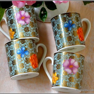 Peut inclure: Quatre tasses décoratives en céramique avec des anses blanches. Les tasses ont un fond bleu clair avec des motifs floraux dorés et des motifs de fleurs colorées. Chaque tasse a une couleur de fleur différente : rose, bleu, orange et jaune. Les bords sont dorés.