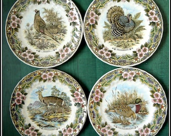May include: Set of four decorative ceramic plates featuring a pheasant, wild turkey, fallow deer, and mallard duck. Each plate has a white background with a green and brown floral border.
