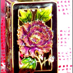 May include: A black and gold decorative bottle with a floral design. The bottle features a purple flower with gold accents and green leaves. The bottle is sitting on a white surface.
