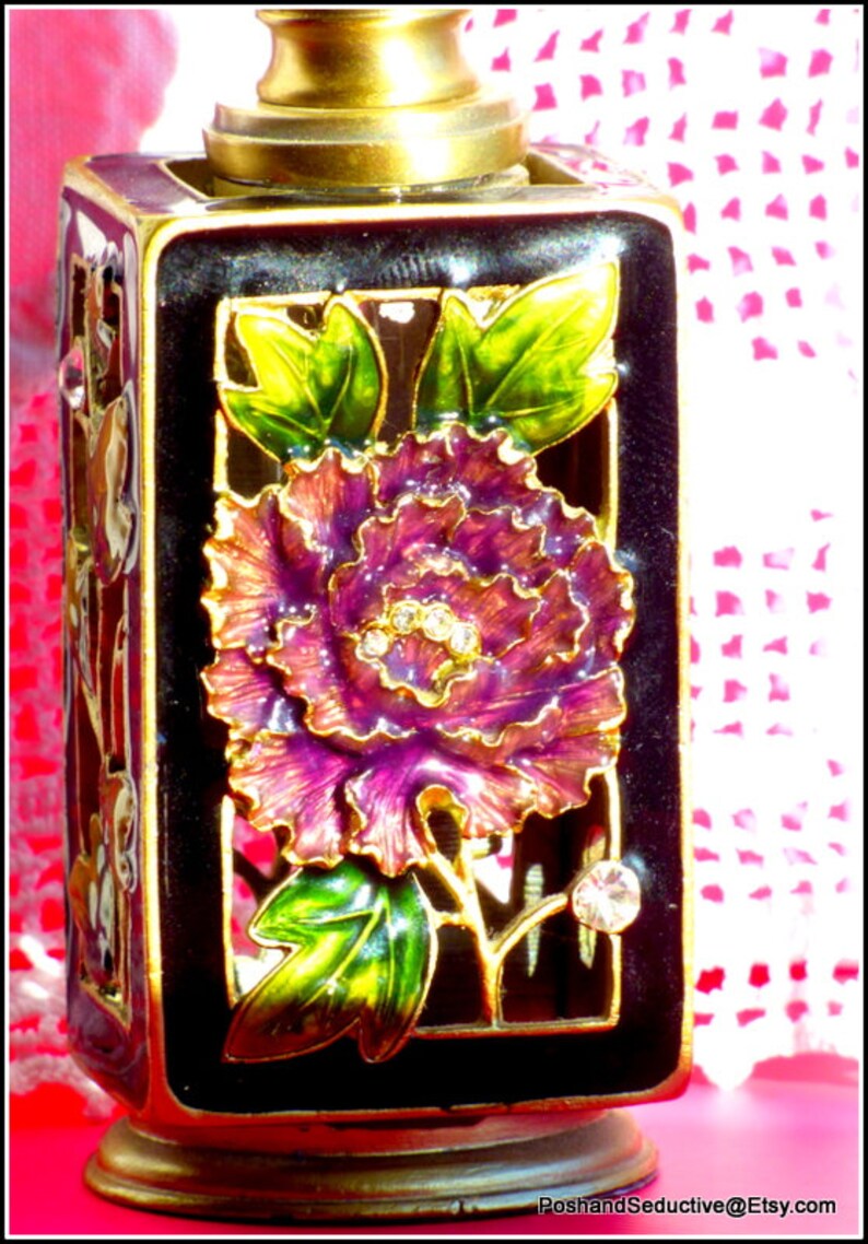 May include: A black and gold decorative bottle with a floral design. The bottle features a purple flower with gold accents and green leaves. The bottle is sitting on a white surface.
