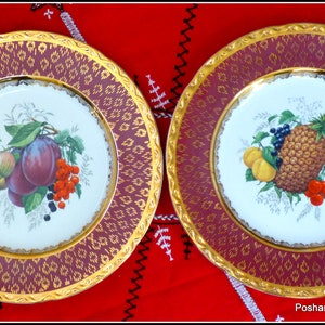 May include: Two decorative plates with gold trim and a burgundy background. One plate features a painted image of plums, grapes, and berries. The other plate features a painted image of a pineapple, cherries, and a peach.