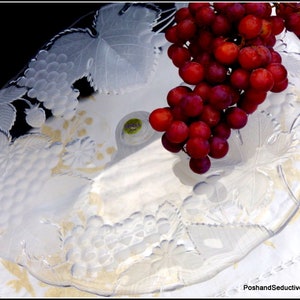 May include: A clear glass serving platter with etched grape and leaf designs. A bunch of ripe, red grapes rests on the platter. The platter is round with a slightly raised edge and sits on a white and gold patterned surface.